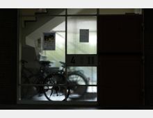 Eine Eingangstür aus Glas mit Strukturglas, hinter der mehrere Fahrräder abgestellt sind. Auf dem Glas ist ein Plakat für ein Sommerfest angebracht, und rechts daneben befindet sich eine Klingel- und Gegensprechanlage mit der Hausnummer „4“.