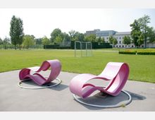 Grüne Parklandschaft mit einer Wiese, auf der sich ein Fußballtor und mehrere Sitzbänke befinden; im Vordergrund stehen zwei geschwungene, modern gestaltete pinke Sitzbänke auf einem gepflasterten Bereich. Im Hintergrund sind Bäume und ein Gebäude zu sehen.