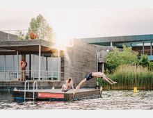 Ein Mann springt von einem Holzsteg ins Wasser, während eine Frau daneben auf dem Steg sitzt. Im Hintergrund ist ein modernes Gebäude mit Holzverkleidung und großen Glasflächen sowie umgebender Vegetation sichtbar.