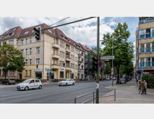 Straßenszene mit Kreuzung in einer Wohngegend, umgeben von gelblichen Mehrfamilienhäusern mit Balkonen und roten Dächern. Im Vordergrund Ampeln, Straßenschilder (u. a. "Gritznerstraße"), parkende und fahrende Autos sowie ein Fahrradgeschäft im Erdgeschoss eines Gebäudes.