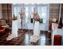 Eleganter Innenraum mit weiß gerahmten Glastüren, gemusterter Tapete und schwarz-braun kariertem Fliesenboden. Im Vordergrund stehen zwei mit weißen Tüchern bedeckte Stehtische mit Blumenarrangements, flankiert von goldenen Kerzenständern und Gruppen kleiner Kerzen in Glasgefäßen.