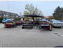 Offene Terrasse mit mehreren Sitzgruppen aus schwarzen Tischen und Stühlen, teilweise mit roten Decken dekoriert. Im Hintergrund sind geparkte Autos, ein Baum und angrenzende Wohnhäuser sichtbar.