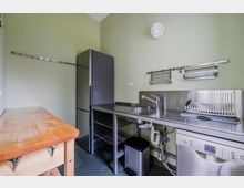 Schlichte Küchenzeile mit grünen Wänden, Edelstahl-Arbeitsplatte und Spülbecken, daneben ein hoher schwarzer Kühlschrank. Unter der Arbeitsplatte stehen eine Spülmaschine und offene Regalböden; an der Wand sind Abtropfgestell und Aufhängeschienen montiert, links ein kleiner Holzarbeitstisch.