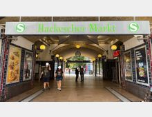 Eingang des Bahnhofs Hackescher Markt in Berlin, mit dem Bahnhofsschild und S-Bahn-Logo über einem mit Rundbögen überdachten Durchgang. Im Inneren sind gelbe Kugelleuchten befestigt, Werbetafeln an den Wänden zu sehen und Menschen gehen durch die Passage.