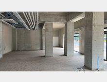 Innenansicht eines unfertigen Gewerberaums mit rohen Betonwänden und -säulen, freiliegenden Kabeln und Lüftungsrohren an der Decke. Große Fensterfronten lassen Tageslicht herein und bieten Ausblick auf die Straße.