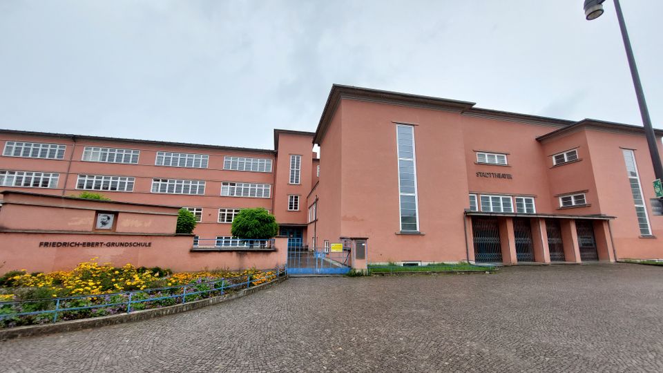 Außenansicht eines Gebäudekomplexes mit orangefarbener Fassade, bestehend aus der "Friedrich-Ebert-Grundschule" und dem "Stadttheater". Links ist ein Schulhof mit Blumenbeeten und rechteckigen Fenstern zu sehen, rechts der Eingangsbereich des Theaters mit hohen Fenstern und Säulenelementen.