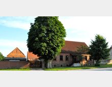 Backsteinbau mit roten Ziegeldächern, umgeben von hohen Bäumen und niedrigen Mauern. Im Vordergrund eine gepflasterte Straße und ein großes Tor, das zu einem Hof führt.