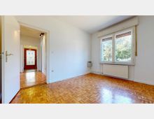 Ein heller Raum mit Parkettboden, einem großen Fenster mit Blick ins Grüne und einer weißen Wandgestaltung. Im Hintergrund führt eine offene Tür zu einem Flur mit roter Eingangstür und Glaseinsatz.