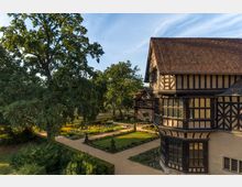 Landschaftsansicht eines Fachwerkgebäudes mit roten Ziegeldächern und verzierten Fenstern, umgeben von einem gepflegten Garten mit symmetrischen Beeten und einem breiten sandfarbenen Weg. Im Hintergrund stehen große Bäume, die den Blick in eine ländliche Umgebung einrahmen.