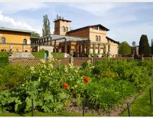 Historische Villa mit gelber Fassade und mediterranen Elementen, flankiert von einem Pergolengang und begrüntem Mauerwerk; im Vordergrund ein gepflegter Garten mit blühenden Pflanzen und roten Mohnblumen.