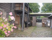 Holzgebäude in Blockbauweise mit rustikalen Details, einem außenliegenden Treppenaufgang und blühenden Hortensien im Vordergrund. Der gepflasterte Innenhof führt durch ein Tor in eine von Bäumen gesäumte Umgebung.