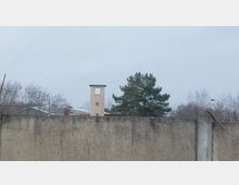 Eine alte Betonmauer mit Metallstreben im Vordergrund; dahinter ein heller Wach- oder Beobachtungsturm und einige Bäume, unter einem grauen Himmel.
