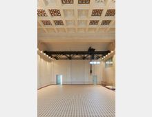 Großer, leerer Innenraum mit heller Kachelbodenfläche im Schachbrettmuster und einer hohen Kassettendecke mit dekorativen Öffnungen. An den Wänden befinden sich große Glasfenster sowie eine Metallkonstruktion mit Kette, die auf industrielle Nutzung hinweist.