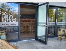 Eingangsbereich einer modernen Weinbar mit großen Glasfronten und geöffneten Glastüren, beschriftet mit „NOR – not only riesling“ und „OPEN“. Durch die Türen sind eine lange Bar, hohe Holzhocker und Regale mit Flaschen im Innenraum sichtbar.