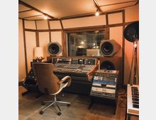 Ein Tonstudio mit einem hölzernen Mischpultschreibtisch, einem großen analogen Mischpult, zwei Studiomonitoren und Racks mit Audioequipment. Im Raum stehen ein Drehstuhl, eine Lampe und ein Mikrofonständer, und durch ein großes Fenster ist ein angrenzender Aufnahmeraum sichtbar.