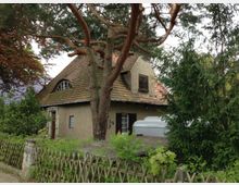 Ein kleines Haus mit schlichter Putzfassade und einem markanten, welligen Ziegeldach, umgeben von Bäumen, Sträuchern und einem Holzzaun. Vor dem Haus steht ein großer Nadelbaum und ein grauer Anhänger ist teilweise sichtbar.