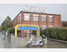 Backstein-Gebäude mit der Aufschrift "OSRAM" auf dem Dach, davor der Eingang zur U-Bahn-Station Paulsternstraße mit einem gelben Eingangsbogen und einem blauen Stationsschild. Im Vordergrund steht ein Motorroller, und Fahrräder sind an einem Geländer abgestellt.