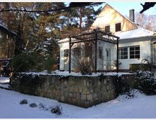 Schnee bedecktes Anwesen mit einem hellen, einstöckigen Haus, das ein Giebeldach und eine Holzfassade an der oberen Etage hat. Im Vordergrund befindet sich eine Terrasse mit einer Pergola auf einer Natursteinmauer, umgeben von Bäumen und winterlicher Vegetation.
