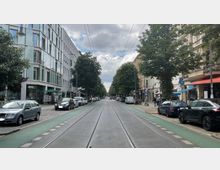 Straßenansicht einer städtischen Allee mit Straßenbahnschienen in der Mitte, flankiert von Bäumen, parkenden Autos und mehrstöckigen Gebäuden mit Geschäften und Restaurants. Der Himmel ist bewölkt, und die Straße ist mit Radwegen auf beiden Seiten markiert.