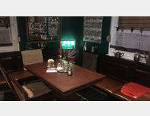 Gemütliche Sitzecke in einem Raum mit dunkler Holzvertäfelung und Dekoration im Retro-Stil, darunter ein Coca-Cola-Spiegel, eine britische Flagge und Vintage-Metallgefäße. Auf dem Holztisch stehen Kerzenhalter, Glasgefäße und kleine Dekorationen, mit Kissen an den Sitzbänken.