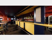 Ein Innenraum einer Bar mit langer Theke in Gelb und Dunkelblau. Im Hintergrund befinden sich Regale mit Flaschen und Gläsern sowie vereinzelt Stehtische und Barhocker auf einem gemusterten Teppichboden.