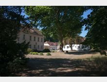 Hof mit historischen Gebäuden, darunter ein großes, helles Haus mit Sprossenfenstern und ein kleineres Gebäude mit roten Ziegeldächern, umgeben von Bäumen und einem offenen, grasbedeckten Bereich. Im Zentrum des Bildes steht ein großer, alter Baum.