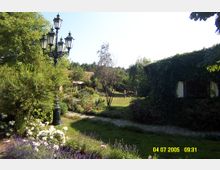 Grüne Parklandschaft mit gepflastertem Weg, bunten Blumenbeeten und einer historischen Straßenlaterne. Rechts ist eine mit Efeu bewachsene Hausfassade zu sehen, im Hintergrund stehen Bäume und eine Wiese.