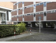 Backsteingebäude mit weißen Fensterrahmen und Fensterbändern, deren unterer Bereich auf Pfeilern über einem Parkplatz schwebt. Im Vordergrund befinden sich ein niedriger Heckenzaun, eine gepflasterte Gehwegfläche und einige rot-weiß markierte Poller.