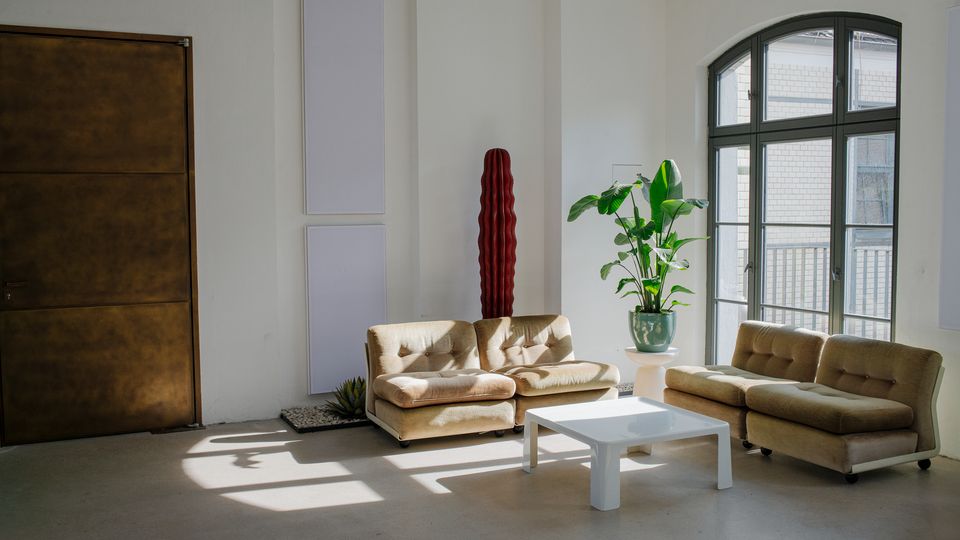 Heller, minimalistischer Lounge- oder Aufenthaltsraum mit weißen Wänden, zwei beigefarbenen Sofas und einem niedrigen weißen Couchtisch auf glattem Betonboden. Ein großes bogenförmiges Fenster mit schwarzem Rahmen lässt viel Tageslicht herein; daneben stehen Topfpflanzen und schlanke Wandpaneele, links eine große braune Tür.