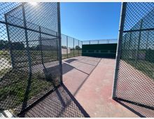 Tennisplatzbereich mit rotem Hartboden, umgeben von hohen Gitterzäunen. Im Hintergrund befindet sich eine grüne Schlagwand mit eingezeichneten Zielmarkierungen.