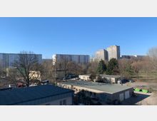 Blick auf eine städtische Landschaft mit mehreren flachen, einfachen Gebäuden im Vordergrund, umgeben von kahlen Bäumen und vereinzeltem Grün. Im Hintergrund stehen mehrere mehrstöckige Wohnblocks vor einem klaren, blauen Himmel.