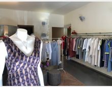 Innenraum eines modernen Bekleidungsgeschäfts mit hellen Wänden. Im Vordergrund steht eine Schaufensterpuppe mit einem gemusterten Kleid, während im Hintergrund Kleidungsstücke auf Kleiderstangen und Kleiderbügeln präsentiert werden.