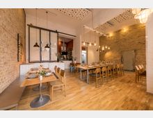 Ein modernes Restaurant oder Veranstaltungsraum mit rustikalem Design, geprägt von sichtbaren Ziegelwänden, hölzernen Esstischen und Stühlen sowie Pendelleuchten. Im Hintergrund befindet sich eine offene Küche mit weißen Fliesen und roten Akzenten.