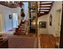 Elegantes Wohnzimmer mit einem Holzfußboden, einer Wendeltreppe aus Holz und einem gläsernen Aufzug in der Mitte. Der Raum ist mit einem großen Orientteppich, einer Wanddekoration, einem Kronleuchter und Bildern geschmückt.