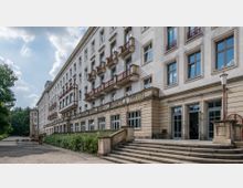 Großes historisches Gebäude mit hell verputzter Fassade, mehreren Reihen von symmetrischen Fenstern und kleinen Balkonen mit roten Geländern. Im Vordergrund befindet sich eine breite Steintreppe, die zu einer Terrasse mit Gartenmöbeln und umgebenem Grünbereich führt.