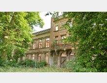 Verfallenes, historisches Gebäude mit sichtbaren Backsteinwänden und großen, leeren Fensteröffnungen, umgeben von dichtem Grün und hohen Bäumen. Die Fassade zeigt verblasste Verzierungen und einen kleinen Balkon mit dekorativen Elementen.