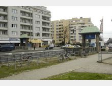 Straßenszene am Theodor-Heuss-Platz in Berlin mit Wohnhäusern im Hintergrund, einem U-Bahn-Eingang mit grünem Dach und Fahrradständern im Vordergrund. Autos und Fußgänger sind auf der mehrspurigen Straße und dem Gehweg zu sehen.