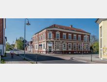 Straßenszene mit einer Kreuzung, umgeben von Gebäuden. Im Zentrum steht ein zweistöckiges Eckhaus im historischen Stil mit roter Klinkerfassade und weißen Fensterrahmen; rechts und links Gehwege, eine Frau fährt mit einem Fahrrad.