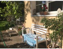 Sonniger Gartenbereich mit einer weißen Holzbank, geschmückt mit einem blauen Tuch, vor einer gelben Hausfassade. Daneben befinden sich Blumentöpfe mit roten Geranien und ein kleiner Baum, während der Hintergrund einen gepflasterten Weg mit Pflanzen und Grünfläche zeigt.