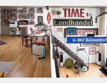 Großzügiger Wohnraum mit Holzfußboden, eingerichtet mit einer Mischung aus Schreibtischbereich und dekorativen Elementen. Die Wand ist mit zahlreichen Schildern, darunter ein U-Bahn-Schild („U-Bhf Bülowstraße“), sowie Bücherregalen und Regalen voller DVDs gestaltet; eine Treppe führt in einen unteren Bereich.