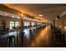 Industrieähnlicher Veranstaltungsraum mit langen Reihen von Stehtischen und Hockern, dezenten Bodenleuchten und Lichterketten an der Decke. Große Bogenfenster lassen Tageslicht herein, und freiliegende Metallsäulen stützen die weiße, gewölbte Decke.