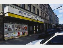 Straßenansicht eines alten Gebäudes mit einem kleinen Kino, das durch ein gelbes Vordach und den Schriftzug "Babylon" gekennzeichnet ist. An der Fassade sind Filmplakate angebracht, und geparkte Autos säumen die Straße.
