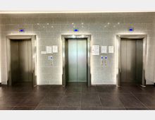 Drei moderne Aufzugstüren in einer Wand aus grauen Fliesen, jeweils mit blauen digitalen Stockwerksanzeigen und Tastenfeldern. Der Boden besteht aus dunklen Fliesen, und an der Wand befinden sich Aushänge und Informationen.