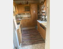 Kleine Küche mit Holzschränken und Arbeitsflächen, einem gefliesten Boden mit einem gestreiften Teppichläufer, Küchengeräten wie einer Kaffeemaschine und einem Mixer sowie Regalen mit Geschirr und Vorratsgläsern.