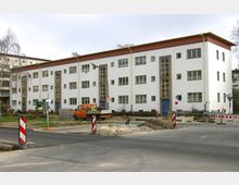 Straßenansicht eines weißen, mehrstöckigen Wohngebäudes im Bauhausstil mit blauen Hauseingängen und vertikalen Fensterelementen. Im Vordergrund befindet sich eine Baustelle mit Straßensperren, Kieshaufen und einem orangefarbenen Transportfahrzeug.