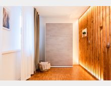 Minimalistisch eingerichteter Raum mit hellem Holzfußboden, einer Schrankwand in Betonoptik in der Mitte und einer dekorativen Holzverkleidung mit indirekter Beleuchtung rechts. Links befinden sich ein Fenster mit lichtdurchlässigen weißen Vorhängen und ein beiges Seitenvorhang.