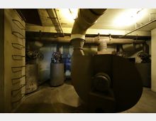 Industrieanlage in einem Kellerraum mit Rohren, Ventilen und großen Metallbehältern. Die Beleuchtung ist schwach, und die Wände sowie die Rohrleitungen wirken abgenutzt.