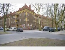 Wohnecke in einer Wohnsiedlung mit mehrstöckigen Gebäuden im Stil der frühen Moderne, gekennzeichnet durch braune Fassaden mit gelben Akzenten, roten Dächern und zahlreichen rechteckigen Fenstern. Vor den Gebäuden stehen parkende Autos, und der Bereich ist von Bäumen und Gehwegen gesäumt.