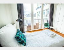 Helles Schlafzimmer mit weißem Bett, buntem Kissen und Blick auf einen Balkon mit Sitzmöbeln. Große Glas-Schiebetüren führen nach draußen, wo moderne Gebäude im Hintergrund zu sehen sind.