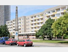 Mehrstöckiges Wohngebäude im Plattenbaustil mit gelben und weißen Balkonfronten, im Vordergrund ein verzierter kleiner Obelisk, geparkte Autos und grüne Bäume.